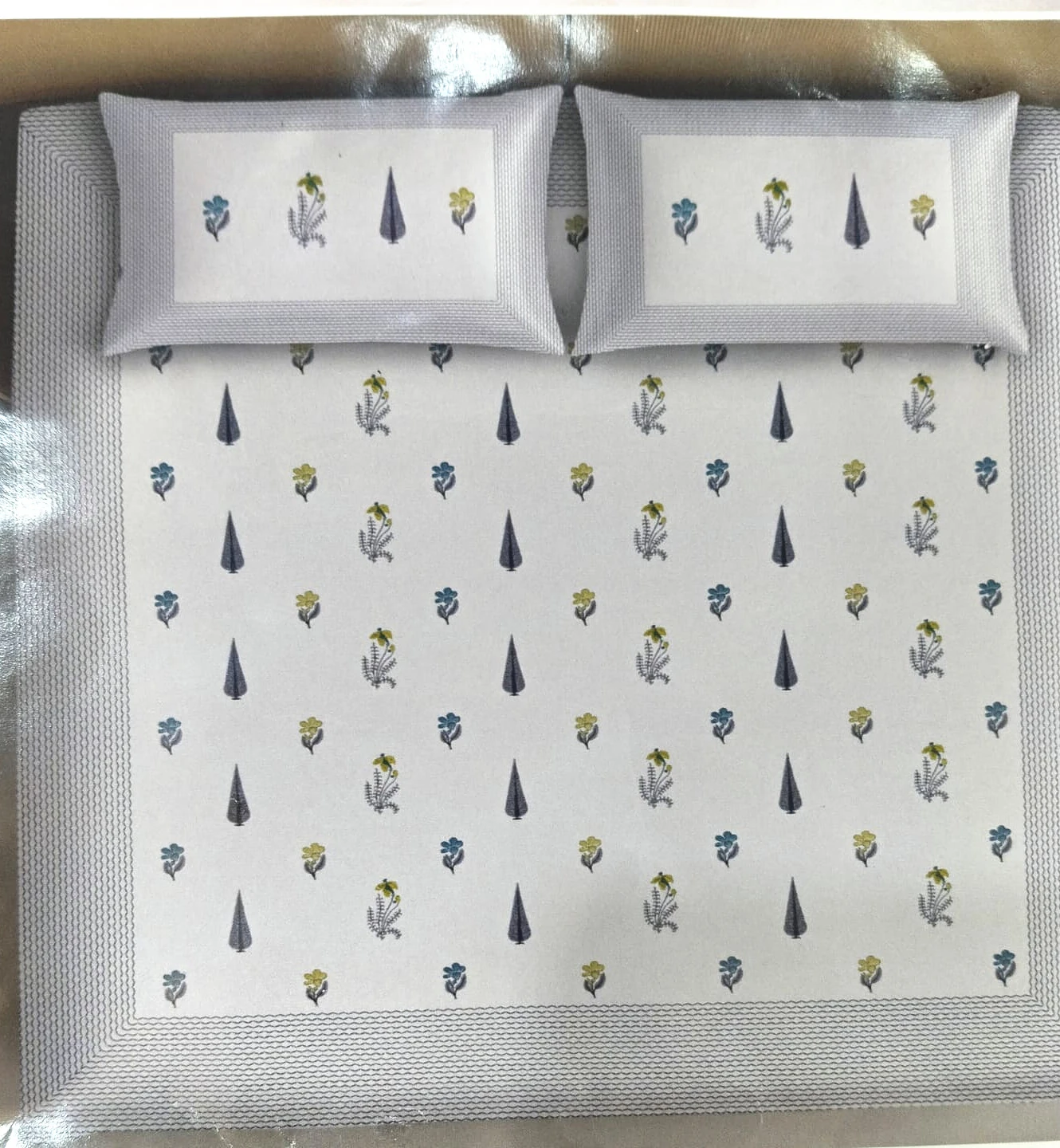 The Wildflower: scattered blue leaves and small yellow flowers on white with a grey border, on a made bed with matching pillowcases.